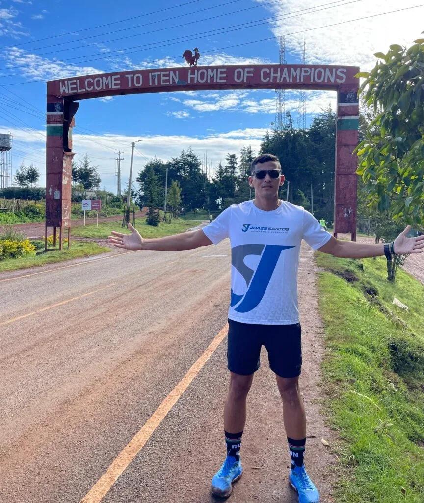 Joaze Santos sob o arco de entrada de Iten, no Quênia, com a inscrição Welcome to Iten — Home of Champions